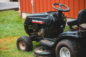Close-up of a black riding lawn mower parked on a lawn next to a red building.